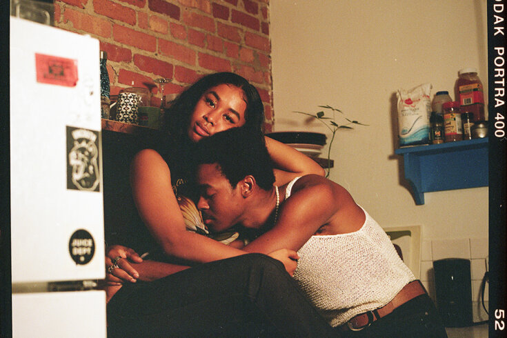 A Black man embraces a Black woman who is sitting on a kitchen countertop. The woman is looking at the camera.